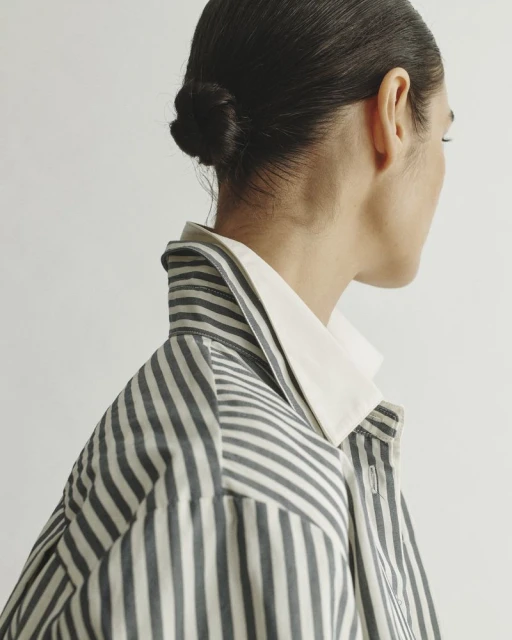 Back view of an adult female with her dark hair in a bun, wearing a vertically striped collared jacket in front of an off-white background.