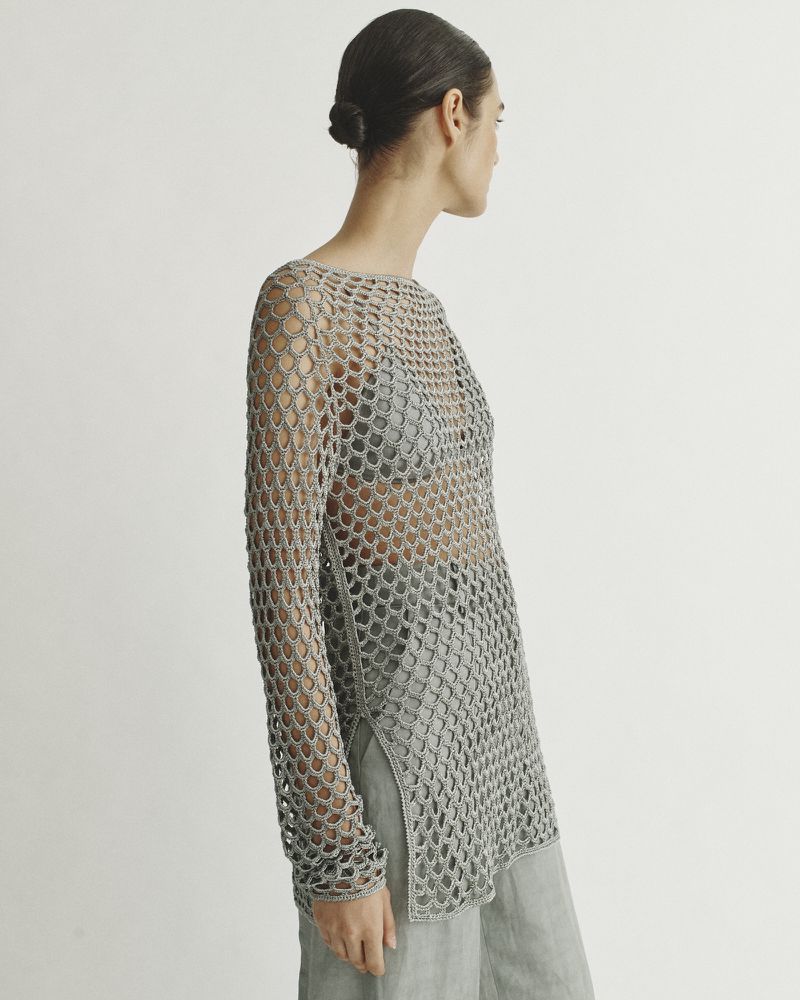 A slender adult woman models a light gray, semi-transparent, net-like, long-sleeve fashion top and matching trousers in a studio setting.