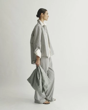 Model wearing striped shirt and matching trousers holding a large grey handbag against a seamless white background.