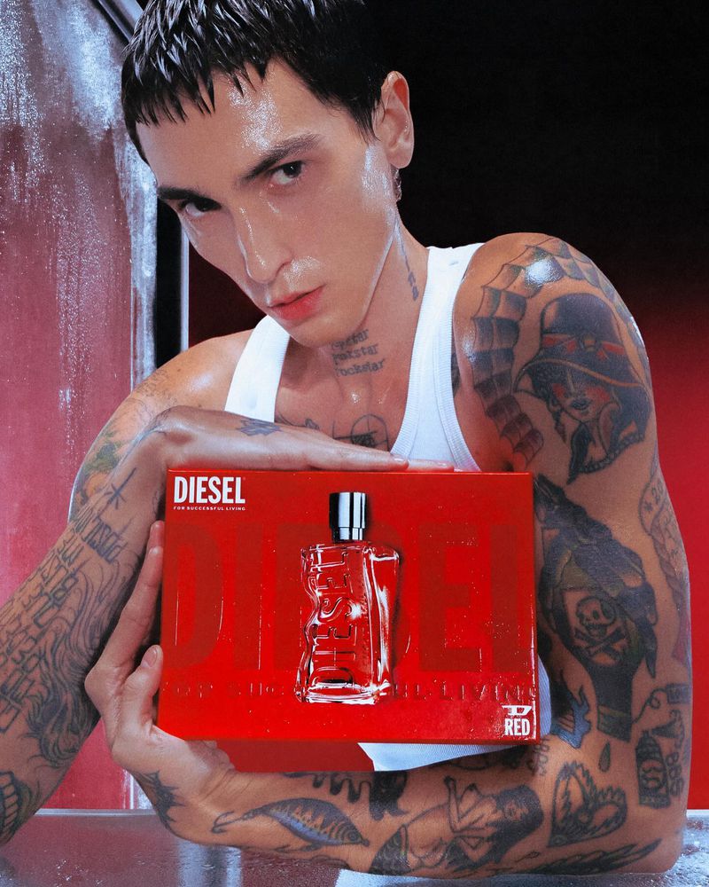 A male model with tattoos poses indoors holding a red Diesel cologne box with a glass cologne bottle, set against a gradient red backdrop.