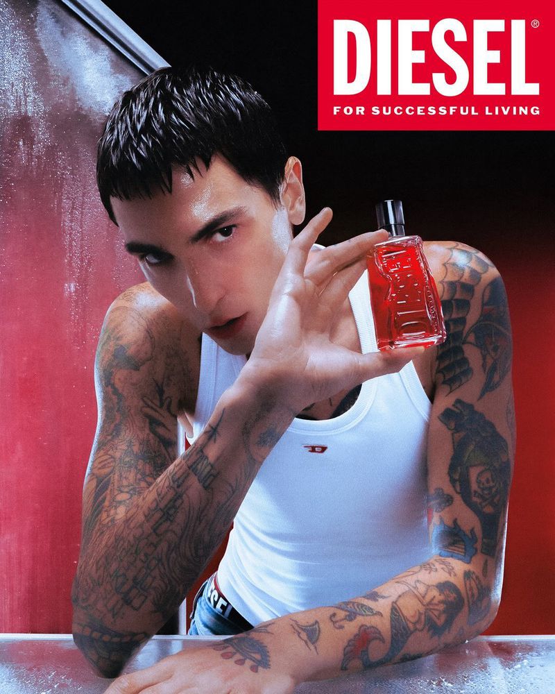 Close-up of a young adult male model with tattoos, holding red Diesel cologne against a red and black backdrop with white trim.