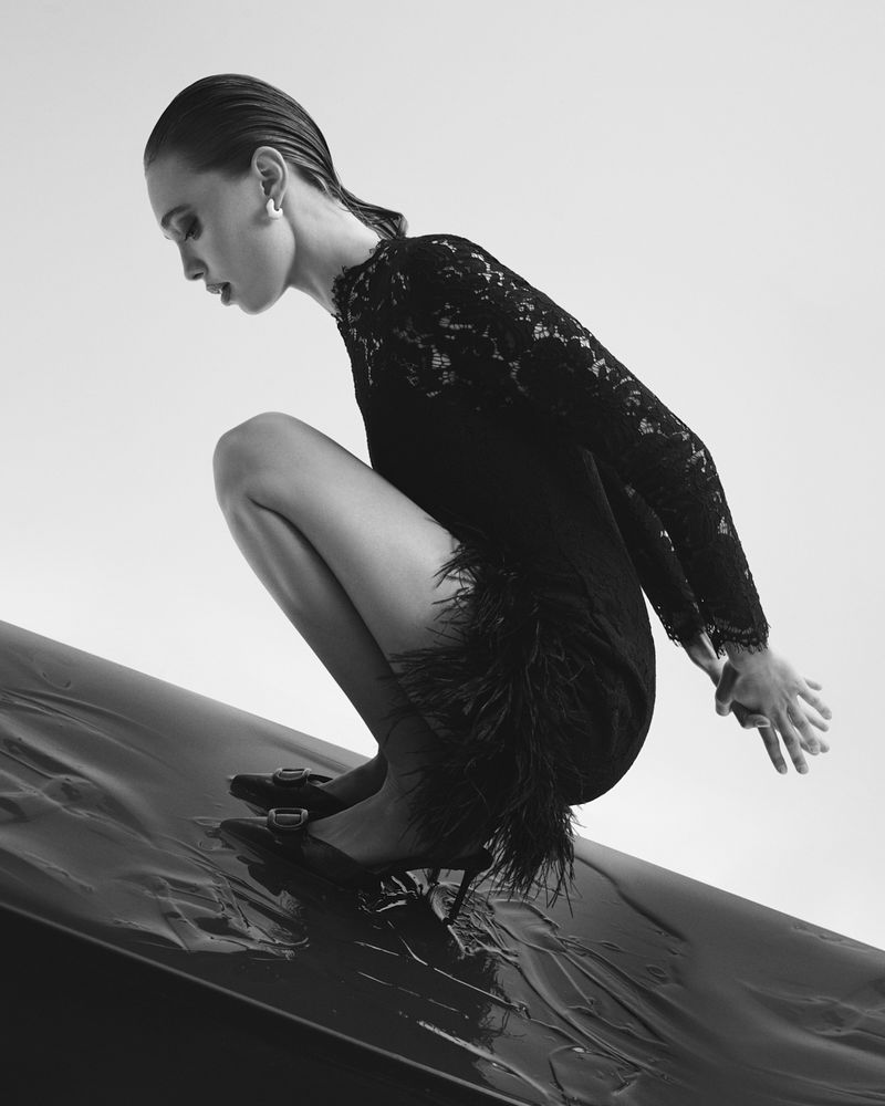 Monochrome fashion shot of a woman crouching in a black lace dress and heels on a black surface, with a light grey background.