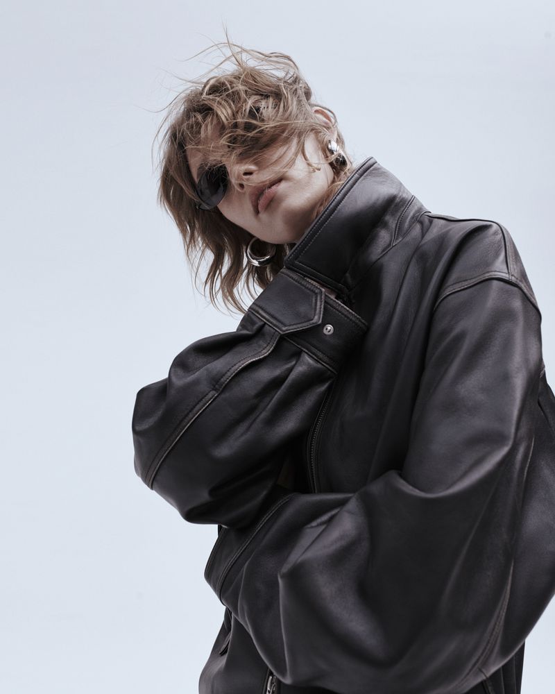 Model wearing dark sunglasses and an oversized black leather jacket against a light blue studio background