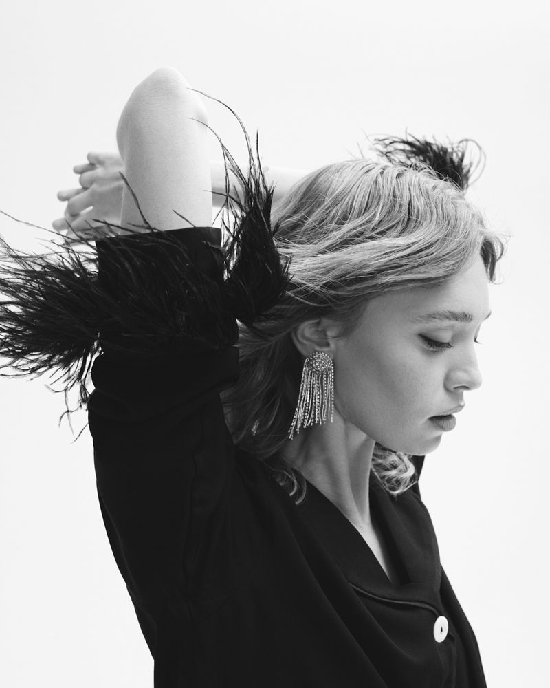 Black and white portrait of a blonde woman in a black top with feather trim, wearing sparkling tassel earrings, against a white backdrop.