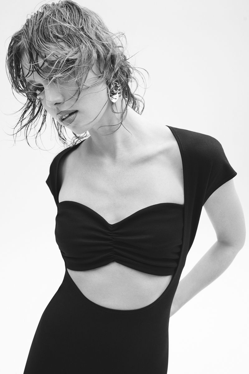 Monochrome portrait of a young adult woman with wet, short hair wearing a black dress in a photo studio with a plain white background.