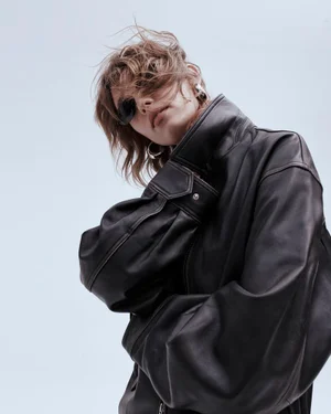 Model wearing dark sunglasses and an oversized black leather jacket against a light blue studio background