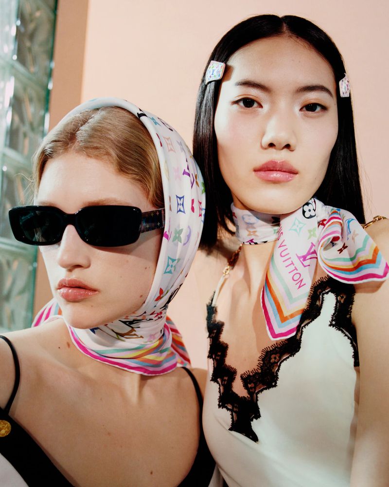 Two women pose indoors wearing headscarves, dresses with lace trim, and sunglasses in a fashion editorial shot with a pink background.