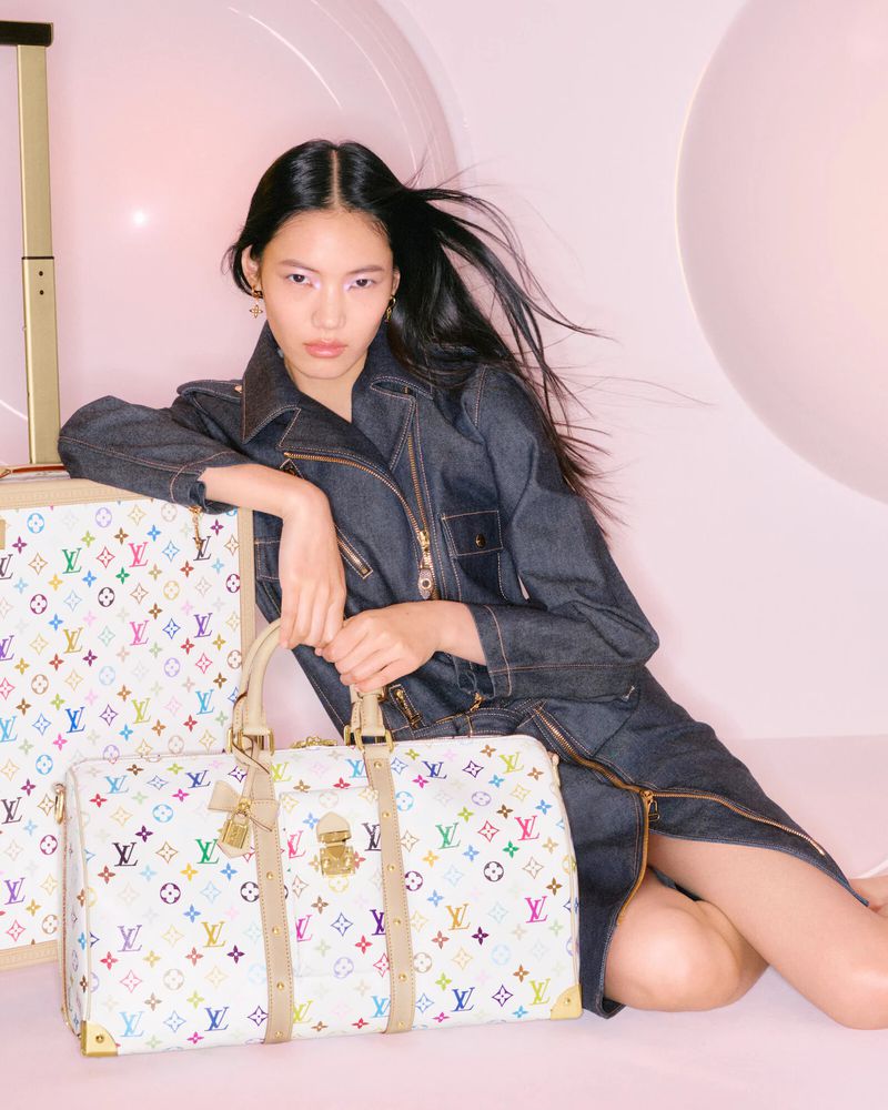 Woman poses with Louis Vuitton luggage. She wears a denim jacket and sits near monogrammed rolling suitcase and shoulder bag.