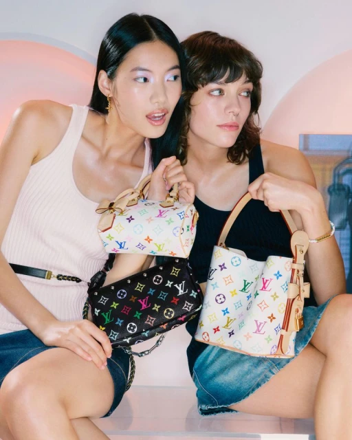 Two women seated indoors, holding colorful Louis Vuitton handbags. The models are wearing casual tops and denim skirts in a studio environment.