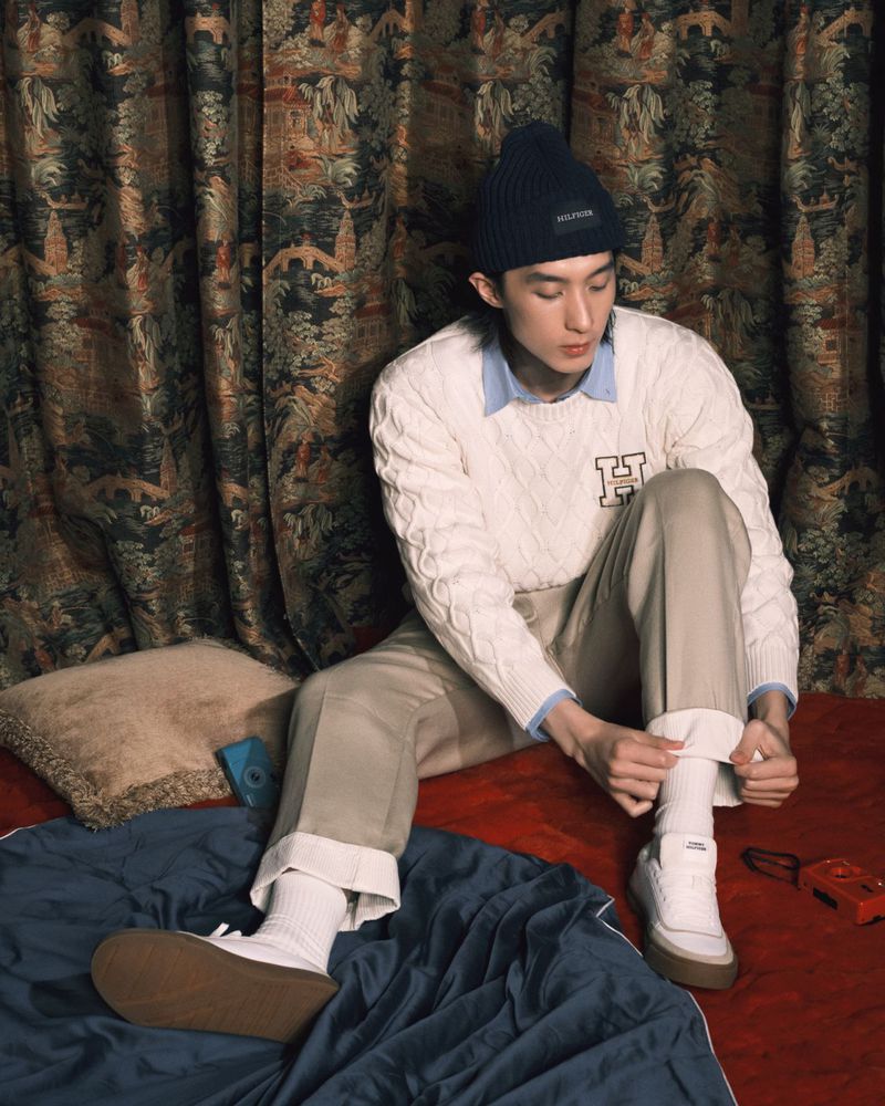 Young adult male in a cream-colored cable knit sweater, light khaki pants, and a dark beanie sits indoors on bedding with a patterned backdrop.
