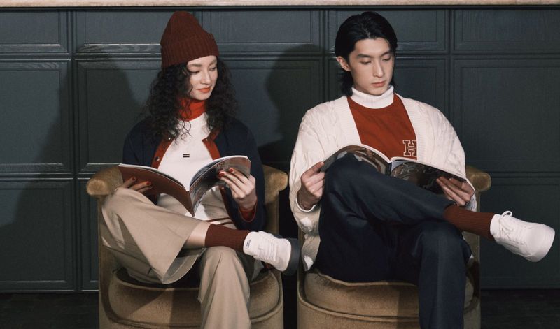 Fashionable young adult couple reading magazines while seated indoors. Woman wears a beanie and man wears a sweater with an 'H' graphic.
