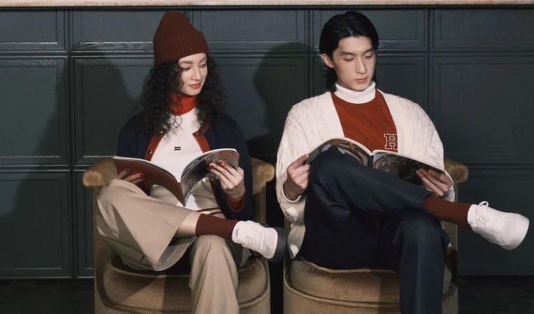 Fashionable young adult couple reading magazines while seated indoors. Woman wears a beanie and man wears a sweater with an 'H' graphic.