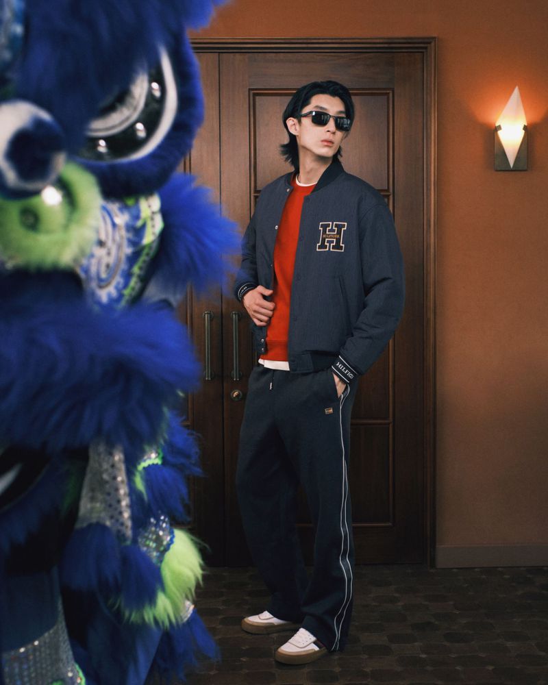 Adult male in fashion shot posing in jacket and track pants next to a blue dragon costume with a light orange wall backdrop.