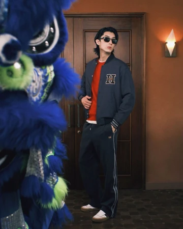 Adult male in fashion shot posing in jacket and track pants next to a blue dragon costume with a light orange wall backdrop.