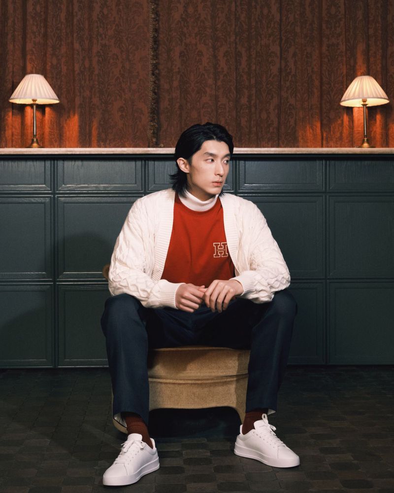 A young adult male sits in a room with a lamp in the background, wearing a red shirt, white cardigan, and white sneakers.