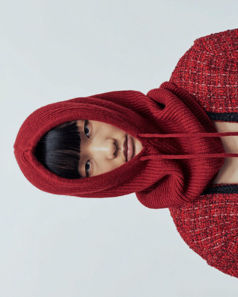 Close-up of a fashionable Asian woman wearing a red knit balaclava-style hood with a matching red jacket against a white backdrop.