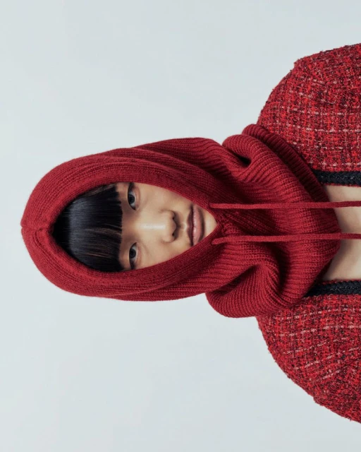 Close-up of a fashionable Asian woman wearing a red knit balaclava-style hood with a matching red jacket against a white backdrop.