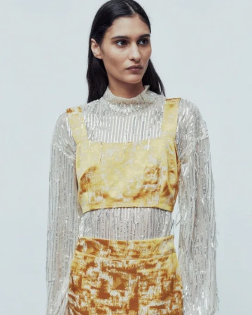 An adult female fashion model poses in a yellow crop top and skirt with a long-sleeved, sheer sequined top against a white background.
