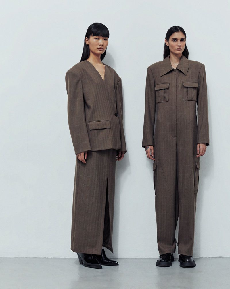Two women stand side-by-side in a studio wearing matching, contemporary brown suit-style outfits and black shoes against a white background.
