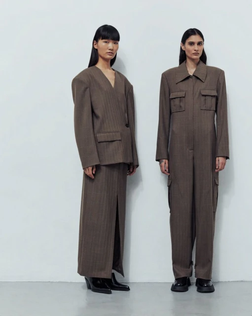 Two women stand side-by-side in a studio wearing matching, contemporary brown suit-style outfits and black shoes against a white background.