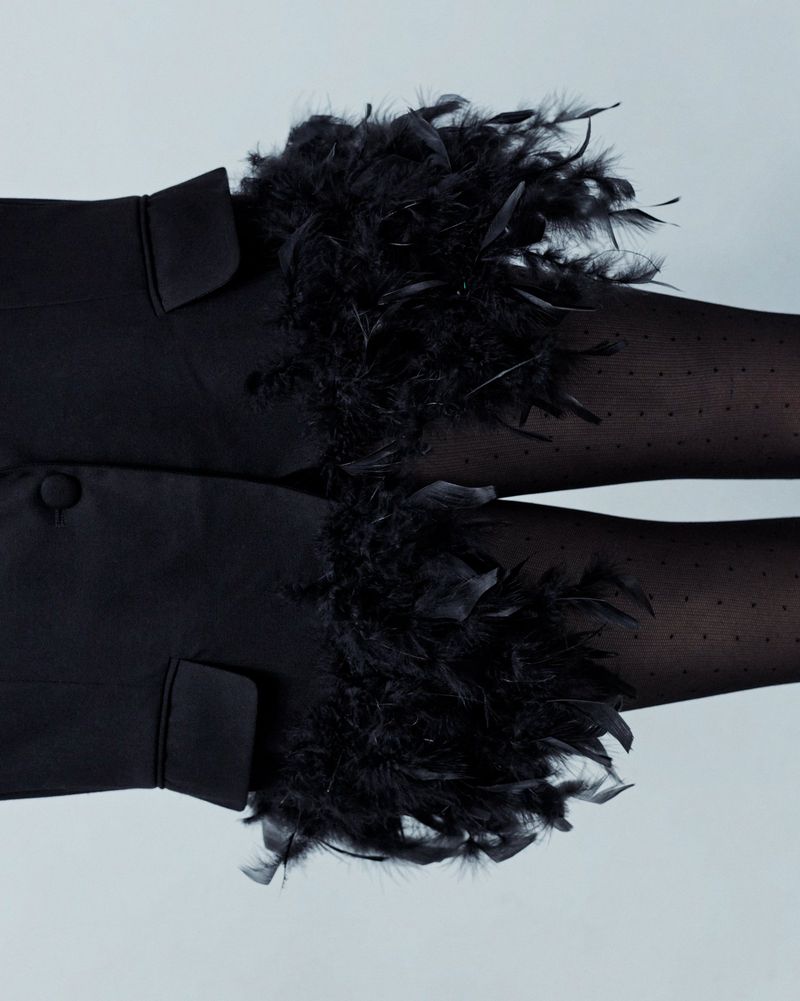 Close-up of a person wearing black dotted tights and a black jacket adorned with fluffy black feather cuffs against a light gray background.