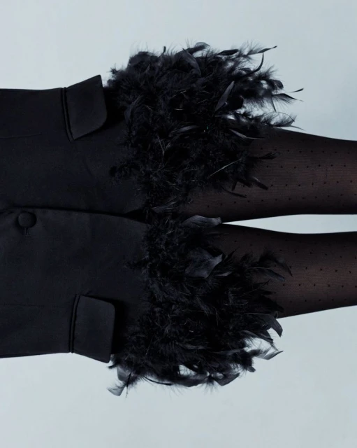 Close-up of a person wearing black dotted tights and a black jacket adorned with fluffy black feather cuffs against a light gray background.