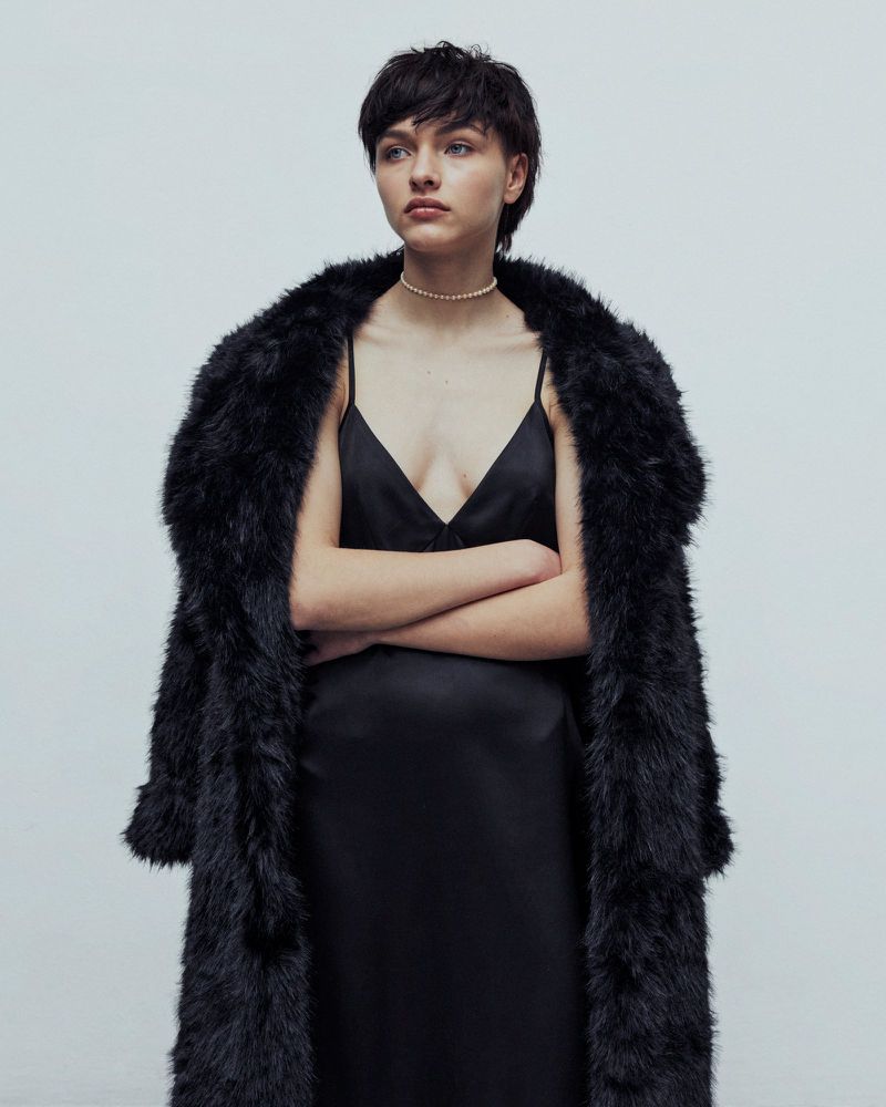 A woman with short dark hair poses indoors, wearing a black slip dress and a long, faux fur coat, against a grey backdrop.