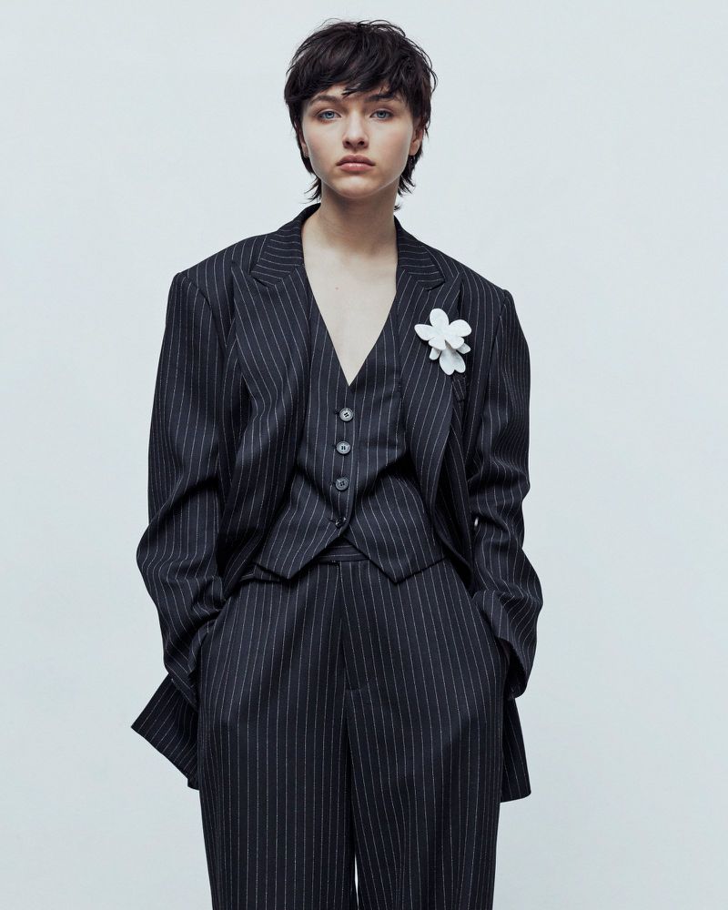 A female model poses in a dark pinstripe three-piece suit with a white flower on the lapel, set against a bright white backdrop in a studio setting.