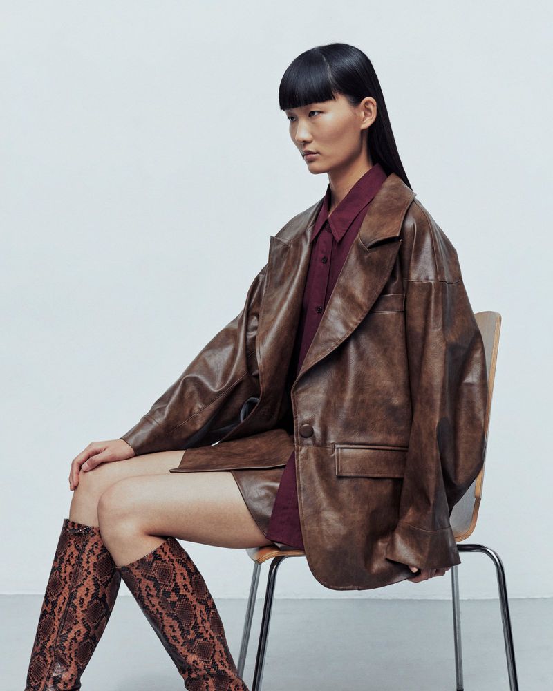Elegant Asian model sits on a chair in a studio wearing a brown leather jacket, burgundy shirt, matching skirt, and snakeskin-print boots.