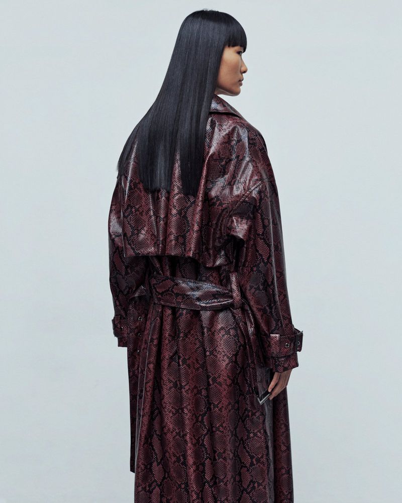 An Asian woman in a snakeskin-patterned trench coat is viewed from behind in a studio, showcasing fashion and style with a modern edge.