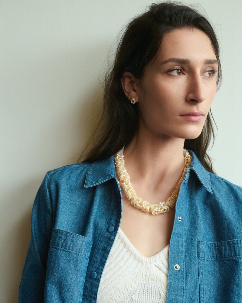 Portrait of a woman wearing a blue denim shirt and a textured white pearl necklace against a plain wall.