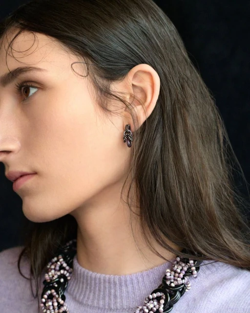 Profile portrait of a young adult female with dark hair wearing beaded jewelry and an earring against a dark background.