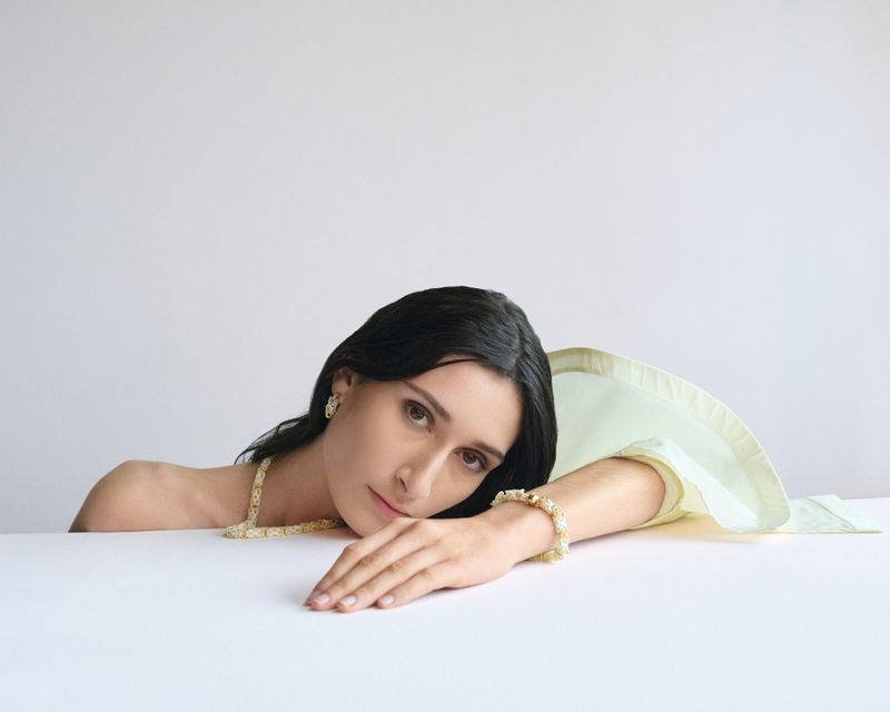 Woman with dark hair poses wearing gold necklace, bracelet, and earrings against a white background, resting her head on her hand.