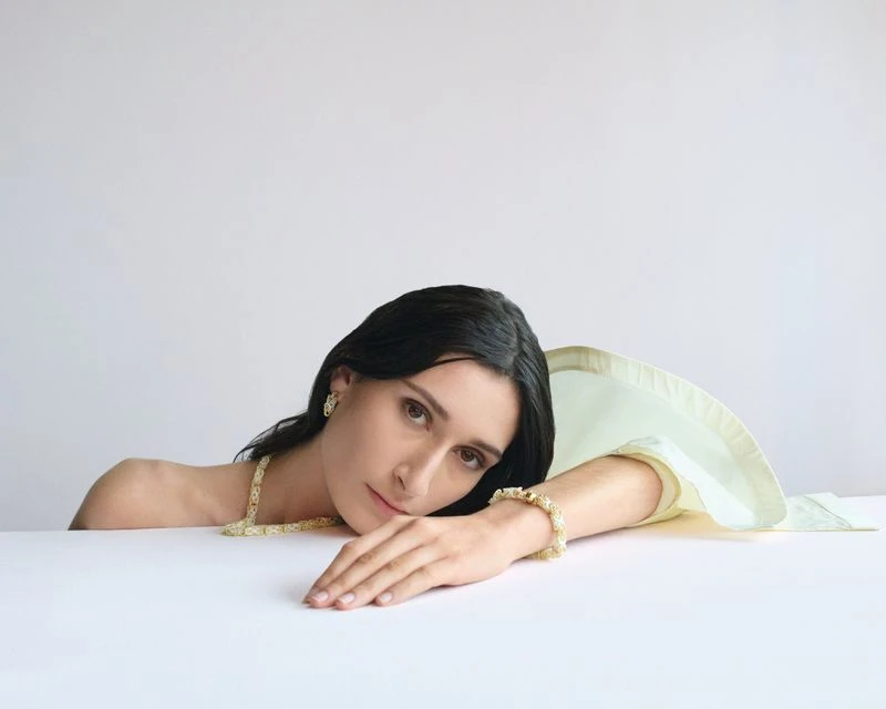 Woman with dark hair poses wearing gold necklace, bracelet, and earrings against a white background, resting her head on her hand.