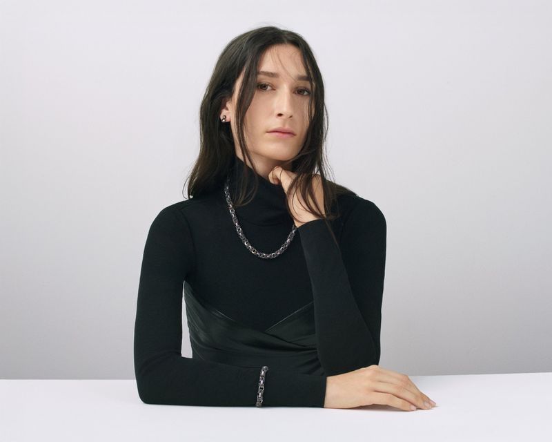 Portrait of a young woman wearing a black turtleneck and jewelry consisting of a necklace and bracelet against a neutral backdrop.