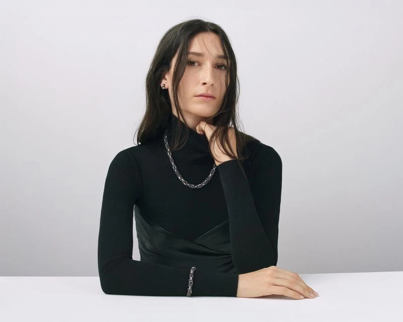 Portrait of a young woman wearing a black turtleneck and jewelry consisting of a necklace and bracelet against a neutral backdrop.