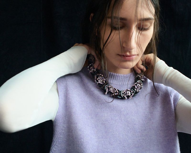 Close-up of a young adult woman with a modern decorative beaded necklace against a dark backdrop, wearing a lilac top.