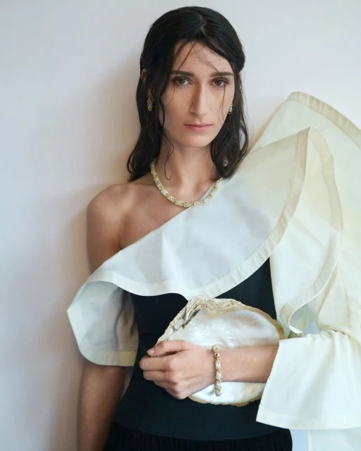 Elegant woman posing indoors wearing a black dress and cream-colored jewelry with a matching clutch, against a light grey background.