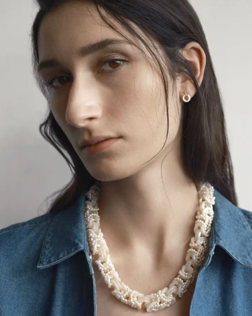 Close-up portrait of a woman wearing a heavy white pearl necklace and matching earrings, styled in a blue denim shirt against a plain wall.