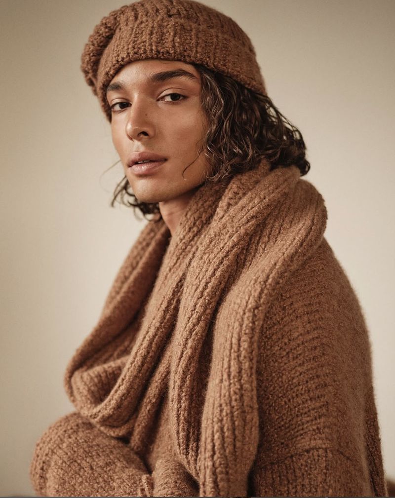 Portrait of a person with long, wavy hair wearing a brown knit hat and a matching oversized knit sweater or scarf, indoors.