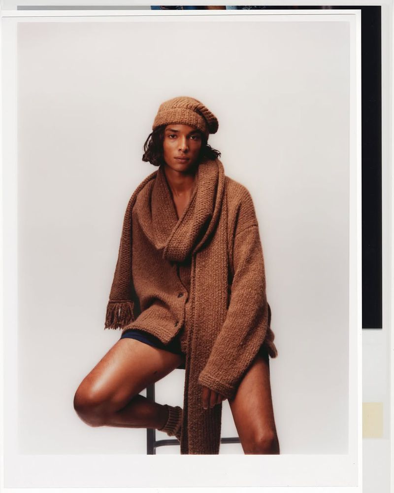 An androgynous person poses in a warm light-toned photo studio, clad in a knit hat, scarf, and cardigan, seated on a stool with legs crossed.