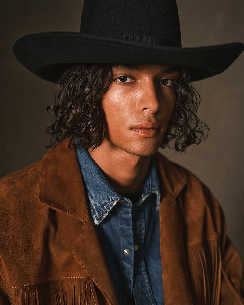 Close-up portrait of a man wearing a black cowboy hat and a fringed suede jacket, posing indoors against a neutral backdrop in soft lighting.