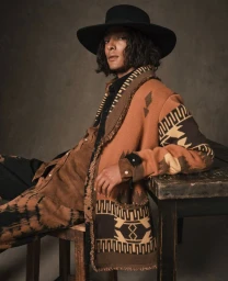 Model Djairo Mulder wearing an patterned knit cardigan and wide black hat seated for the Alanui FW24 campaign by Giampaolo Sgura.