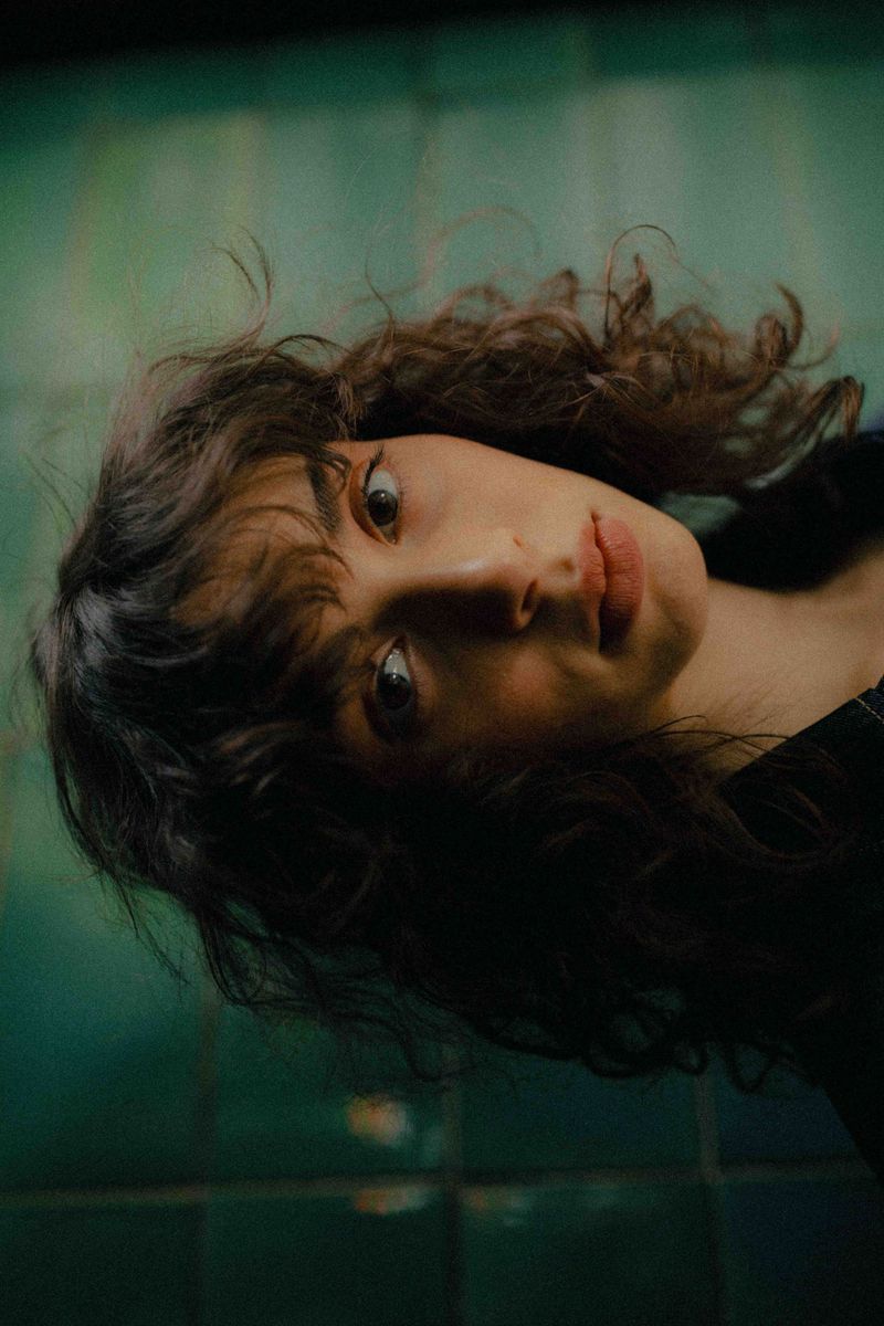 Close-up portrait of a young adult female with curly dark hair looking at the camera against a green tile background indoors.