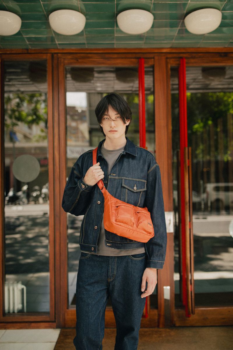 A young Asian man wearing a denim jacket and jeans poses in front of a glass-paned door, sporting an orange crossbody bag for a stylish look.
