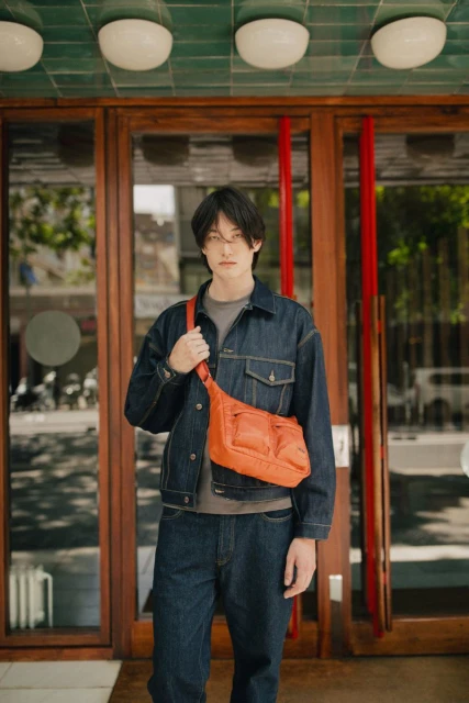 A young Asian man wearing a denim jacket and jeans poses in front of a glass-paned door, sporting an orange crossbody bag for a stylish look.