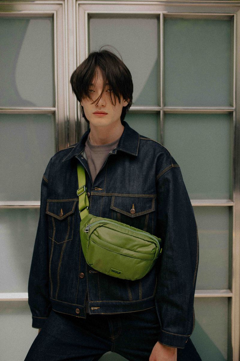 Person poses wearing a denim jacket and carrying a green fanny pack in front of a window with translucent glass panels.