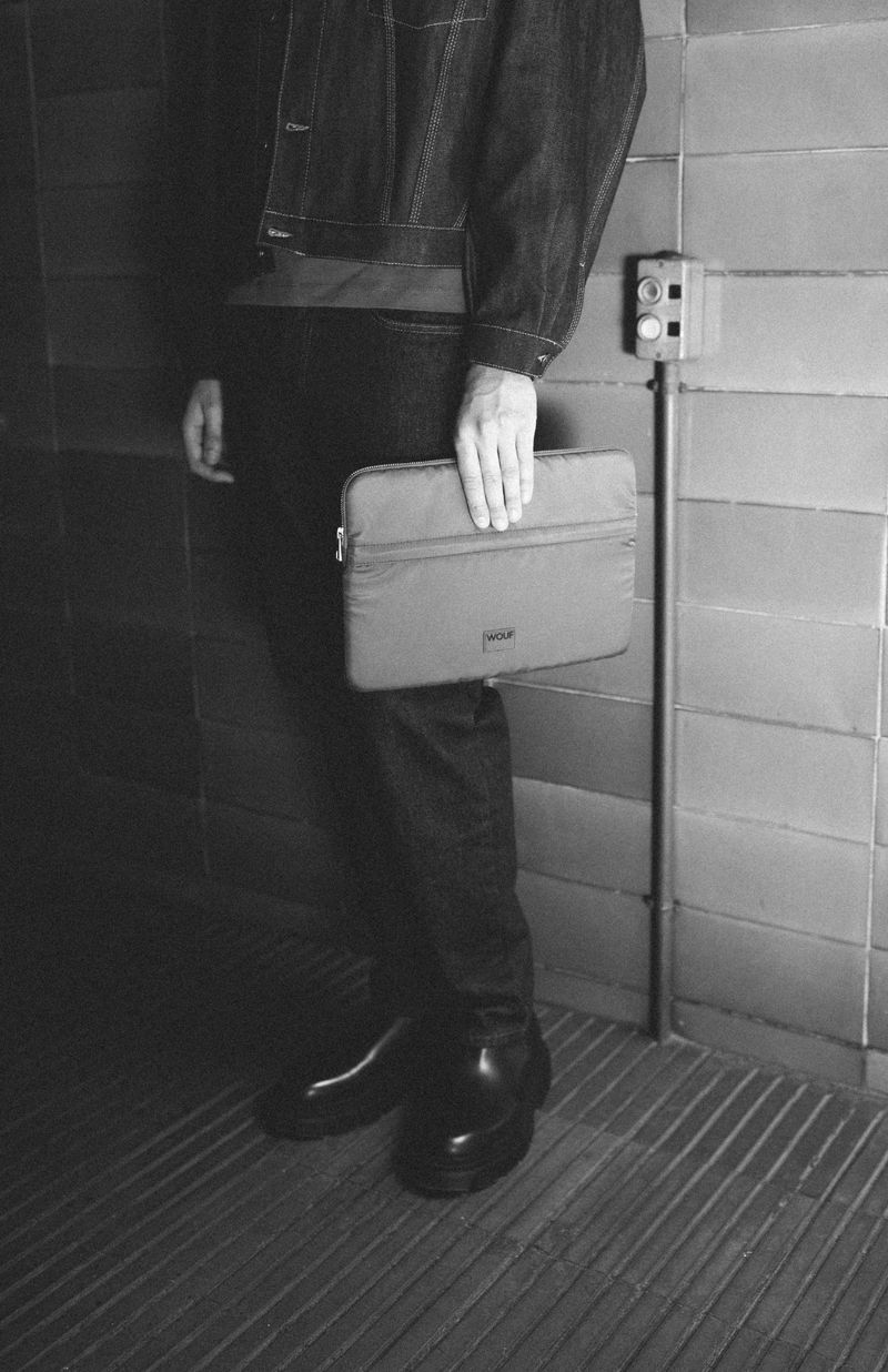 Person in denim jacket holding a laptop case against a tiled wall, creating a stylish and practical fashion product shot.