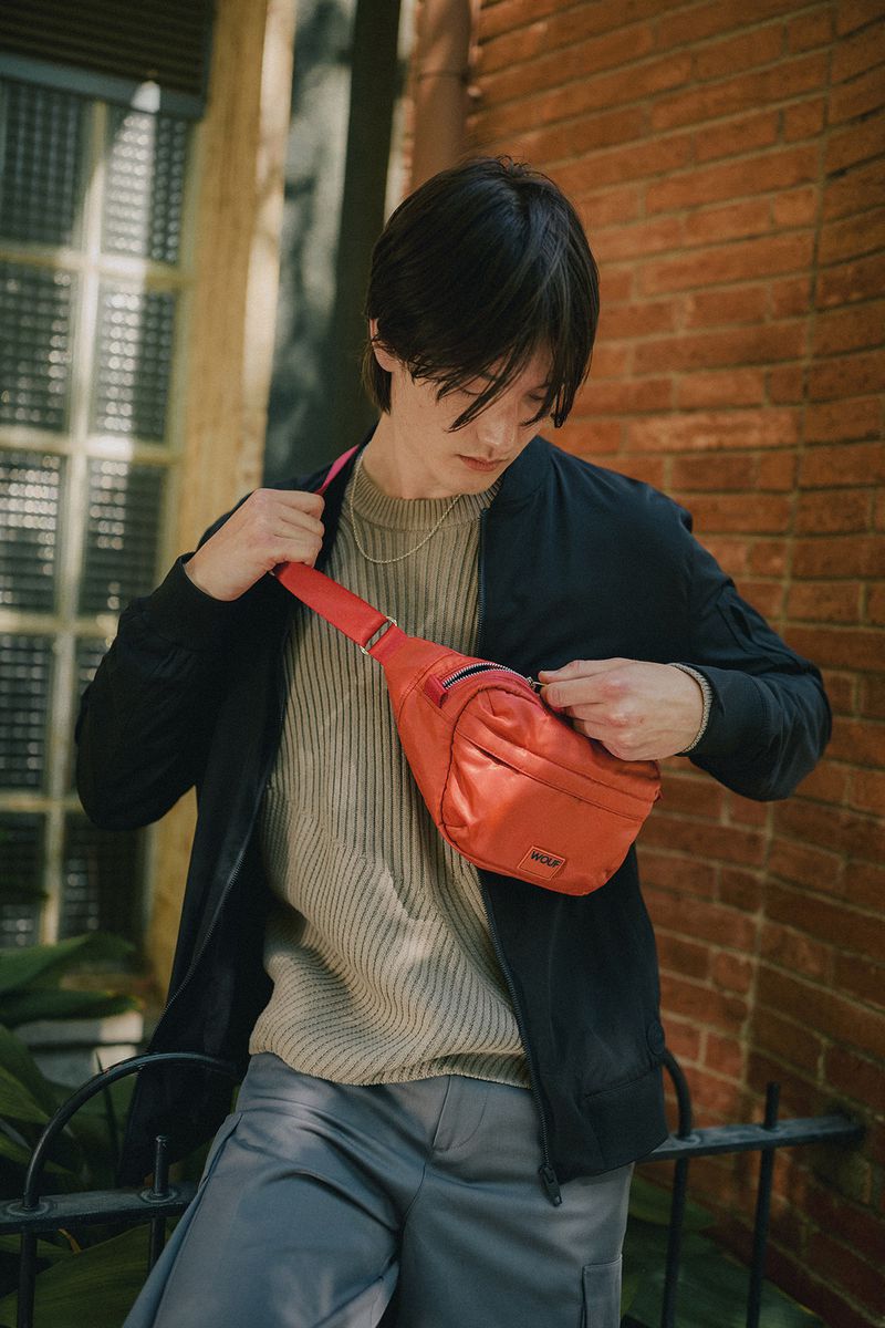 Young man outdoors with black bomber jacket and grey trousers wearing a red fanny pack, standing against a brick wall.