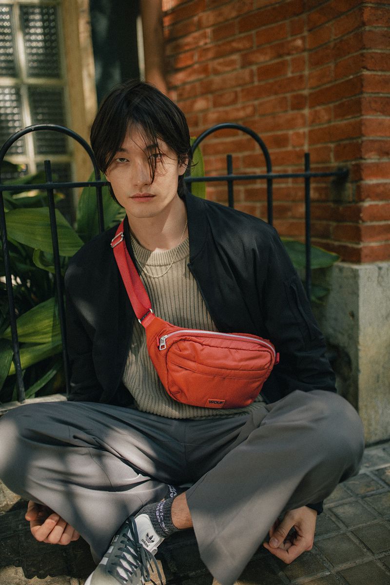 A stylish person sits outdoors with a red belt bag, gray pants, black jacket, and Adidas shoes, posing against a brick wall.